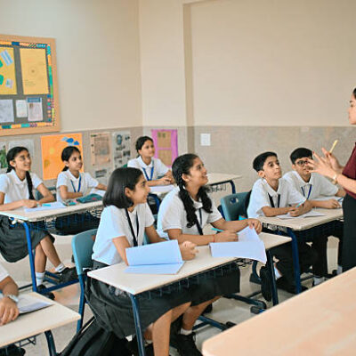 Female Indian teacher standing at the front of a classroom, explaining a lesson to a group of attentive middle school students. Boys and girls sit at their desks with notebooks open, listening, asking questions, and engaging in the discussion, showing active learning, education, and positive teacher–student interaction in an Indian school setting.