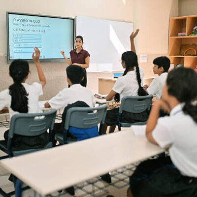 Group of Indian school students in uniform attending a math class, raising their hands to answer a question displayed on the smart screen. Female teacher stands near the board conducting a quick classroom quiz on quadratic equations. Concept of active learning, STEM education, student participation, and modern classroom environment in India.