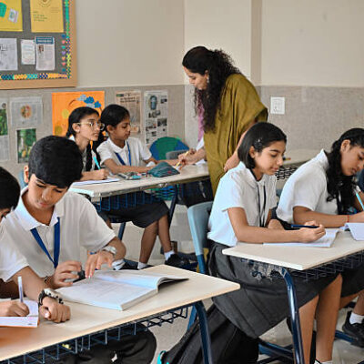 A group of Indian middle-school students sitting at their desks and working in notebooks while a female teacher in a saree walks around the classroom, assisting a girl with her lesson.