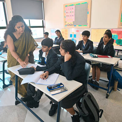 A teacher in a sari bends over a front-row desk, to explain something to two students. The students listen closely with interest. The classroom is bright, with large windows, bulletin boards, and school bags beside the desks, capturing a moment of hands-on guidance during a lesson.